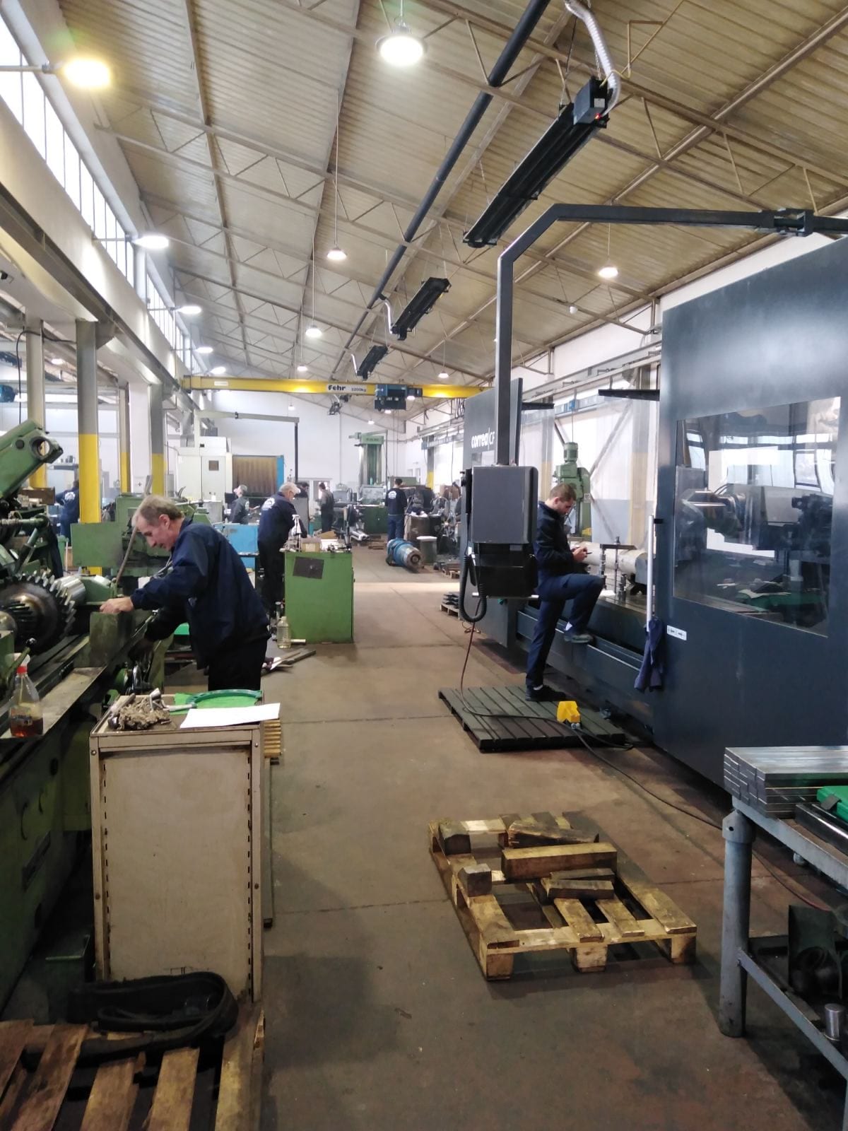Mašinska hala u radu: radnici za konvencionalnim i CNC mašinama, desno velika CNC mašina, iznad žuta mosna dizalica